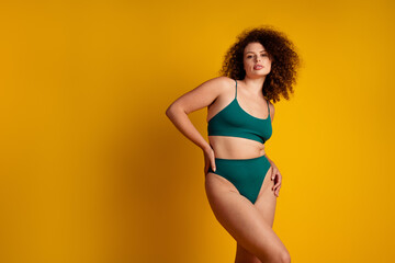 Confident young woman with curvy body in green lingerie posing against vibrant yellow background radiating beauty