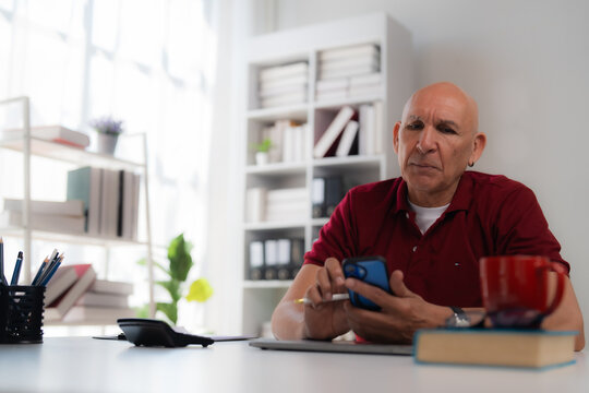 Focused mature businessman using smartphone and digital pen while working at desk in bright modern office, with books, pencils, phone and laptop on table