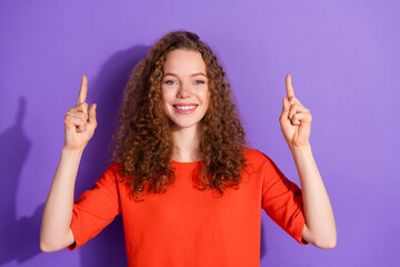 Smiling young woman in casual orange attire pointing upwards with both hands against a vibrant...