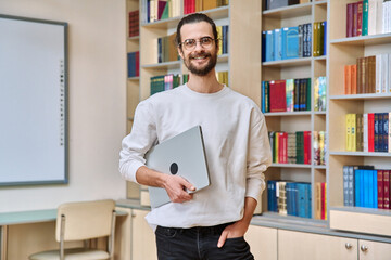 Portrait of young male teacher, mentor, adult student holding laptop, inside library classroom