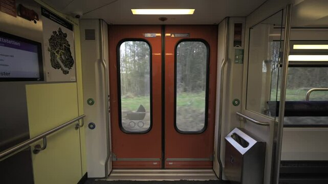 Interior of Swiss double-decker train showing orange automatic doors with windows, handrails and digital display. Train moving through countryside, highlighting modern public transport infrastructure