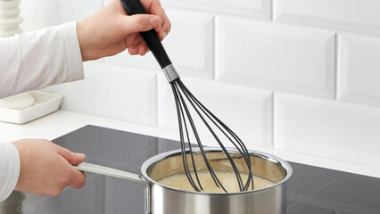 Cooking a creamy sauce in a stainless steel pot using a whisk on a modern stovetop in a bright...