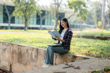 Obraz premium Female student reading book while sitting on wall, green campus landscape behind her