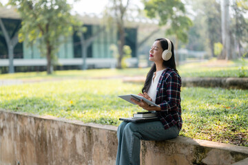 Asian student enjoying music with headphones while using tablet for studying, sitting in a university campus park
