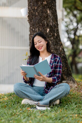 Young woman reading book, resting on grassy ground beneath tree shade, representing learning and...