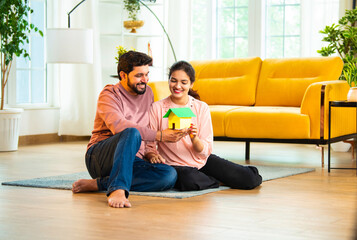 Smiling Asian couple holding colorful paper house model while planning new home in living room