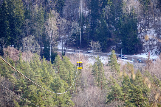 
A yellow aerial tram car moving above a forested landscape with a road and cars in the background.