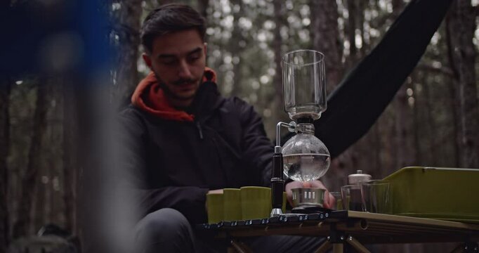 A man makes coffee on a winter picnic in the woods. He uses a syphon coffee maker on a portable table.