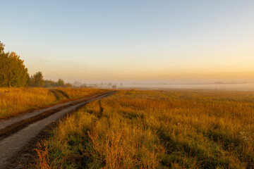 Fototapeta premium Picturesque view of landscape against sky in foggy weather. Warm autumn morning. Picturesque view of road in field against sky during sunrise