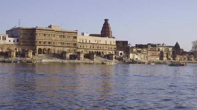 At Keshi Ghat in Vrindavan, the view across the Yamuna River includes people moving along the banks. Boats moving in the water add to the lively scene.