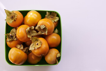 Bunch of ripe persimmons lies in a square green bowl on a white table. Top view