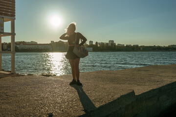 Woman Silhouette Lake Sunset - A lone woman stands on the edge of a lake, her silhouette backlit by the setting sun. The city skyline can be seen in the distance.