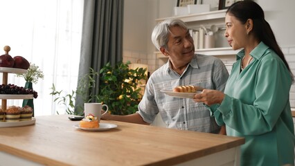 Energetic grandmother holding breakfast and serving to her husband while grandfather sitting at modern kitchen. Smiling elder couple spending time together. Retirement lifestyle concept. Myrmidon.