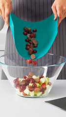 Person pouring sliced grapes from flexible cutting mat into glass bowl.