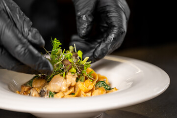 A chef in black gloves plates creamy pasta with mushrooms and greens on a white dish, adding microgreens for garnish. Emphasizes attention to detail and gourmet presentation.