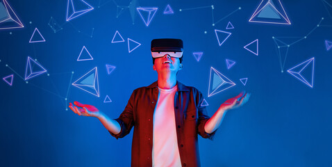 Excited man with casual shirt excited and stands at blue background and looking floating shape. Happy player accessing metaverse and entering visual reality world. Technology innovation. Ingenuity.