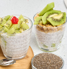A glass of chia seed pudding layered with kiwi and granola, topped with kiwi slices, sits on a wooden board with ingredients around it