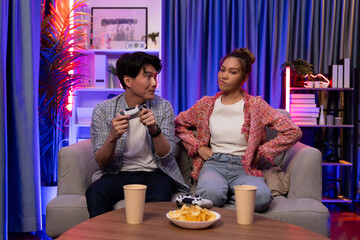 Sitting sofa of happy couple playing video game with joysticks, smiling man playing challenge, raising fist up to be winner the challenge at modern neon blub light living room at home. Infobahn.