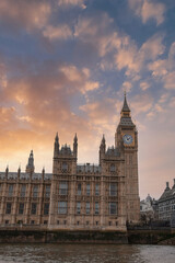 Fototapeta premium Elizabeth Tower, part of the Palace of Westminster, stands against a sunset sky with orange and pink hues. The River Thames reflects the soft evening light.