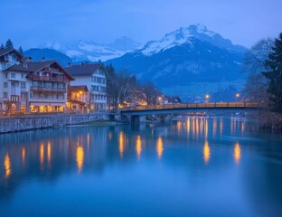 Fototapeta premium Serene Swiss Village at Dusk: Tranquil River, Illuminated Buildings, Majestic Mountains