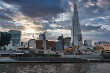 Fototapeta premium HMS Belfast, a historic Royal Navy warship, moored on the River Thames in London, with The Shard and modern office buildings under a partly cloudy sky.