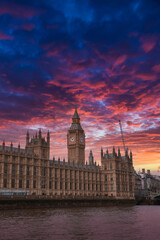 Fototapeta premium Elizabeth Tower, part of the Palace of Westminster, illuminated by a vibrant sunset. The River Thames reflects the colorful sky, with a crane visible nearby.