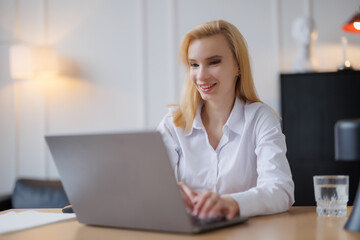 Obraz premium Young woman works on laptop in bright office during daytime with a glass of water nearby and a warm atmosphere
