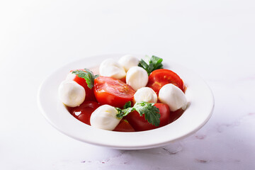 Juicy caprese salad with baby mozzarella cheese, cherry tomatoes and parsley in white ceramic plate on white marble table close up. Healthy diet food concept.