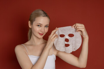 Young attractive asian woman on red background with moisturizing face mask. Skin care concept, skin care cosmetics for skin moisturizing