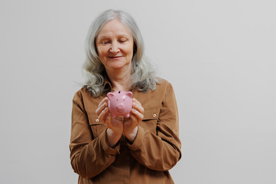 Older woman smiles while holding a pink piggy bank in a neutral-colored room during a financial planning session