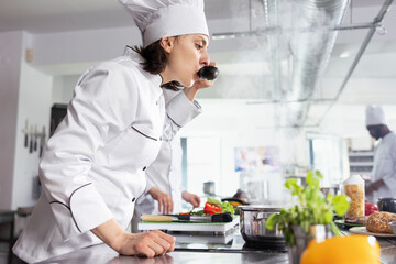 Female chef in uniform leans over the stovetop and tasting a delicious dish with a spoon. Focused...