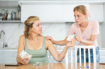 Focused woman received salary counting money at kitchen table while teenage daughter patiently waiting for pocket money..