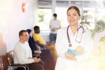 Polite young female therapist in white coat standing in clinic, filling clipboard with medical records