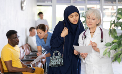 Experienced aged female doctor showing results of medical examination and explaining prescribed treatment to young asian woman wearing traditional muslim dress in lobby of clinic