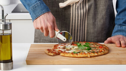 Person uses a pizza cutter to slice a freshly baked pizza on a wooden board in a modern kitchen...