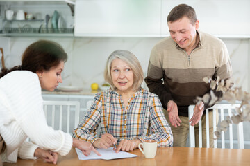 Thoughtful old woman compiling heritage document while middle-aged man and woman competing with...