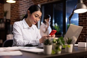 Female employee taking a break at work, using her mobile device at her office desk during her night shift. Young asian manager texting on her smartphone, communicating with remote colleagues.