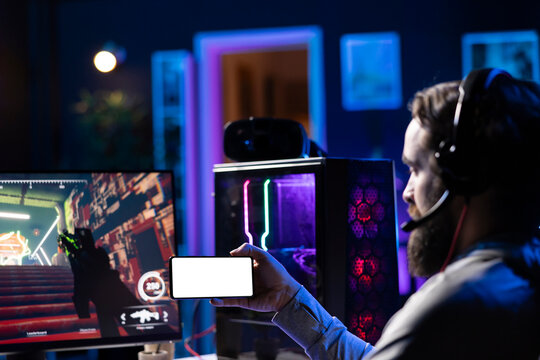 Man watching tutorial on isolated screen mobile hone while playing first person shooter videogame with guns. Gamer learning to play game by looking at online guide on mockup cellphone
