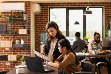 Fototapeta premium Confident female employees concentrating on business analytics, using clipboard to review financial documents. Caucasian woman standing and having a discussion with asian coworker in wheelchair.