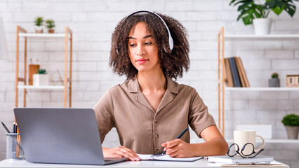 Self-isolation, online education concept. African american millennial student in headphones sit at desk, watch webinar and making notes