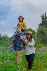 Fototapeta premium Happy family in country side meadow. Child in yellow sweater with stuffed animal on dads shoulders, mom in hat and white shirt. Perfect for family, nature, or lifestyle themes.