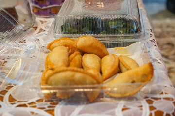 A selection of Malaysian snacks displayed in clear plastic containers, featuring golden-brown karipap (curry puffs) and kuih koci in green banana leaf.