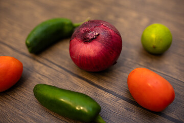 close-up of a red onion, poblano pepper, red tomato, and a lime against a dark wooden surface