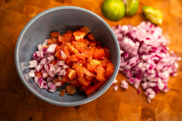 close-up of a gray blue bowl with chopped red onions and chopped red tomatoes on a wooden cutting board with additional chopped red onions and lime slices, Mexican food