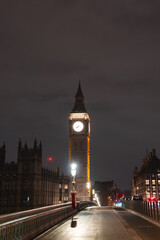 Obraz premium Big Ben's illuminated clock tower shines against a dark sky, with Westminster Bridge in the foreground featuring ornate railings and warm streetlights.
