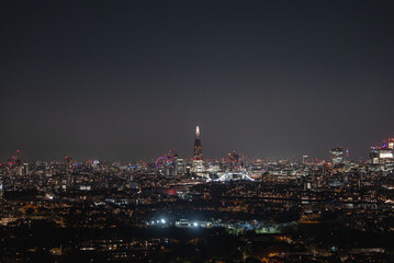 Illuminated Shard skyscraper at night with Tower Bridge spanning the River Thames. The London Eye is visible in the distance under a clear sky.