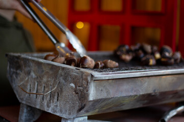 Defocused photo close up. Process of cooking food on grill. Cook roasts chestnut with tongs. Street fair on city holiday: Harvest day, Christmas, Tbilisoba, New Year. Concept of lunch, dinner, snack