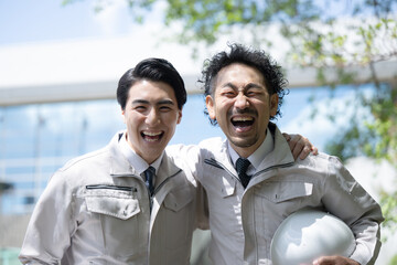 Image of construction management and other workers looking up at the front of a building in work clothes under a blue sky, looking at the camera, shoulders together.