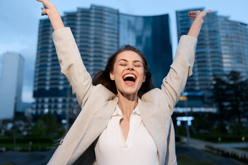 Joyful businesswoman celebrating success outdoors in urban setting, wearing stylish blazer, evokes confidence and positivity with vibrant city backdrop.
