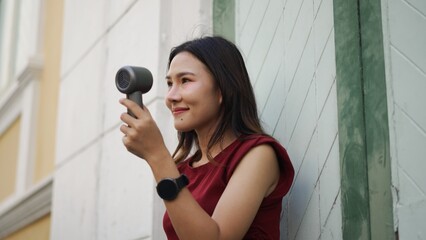 young asian woman using portable handheld fan outside, smiling in casual red outfit with smartwatch, enjoying cool summer breeze, showing modern lifestyle, beauty and relaxation © pixs4u